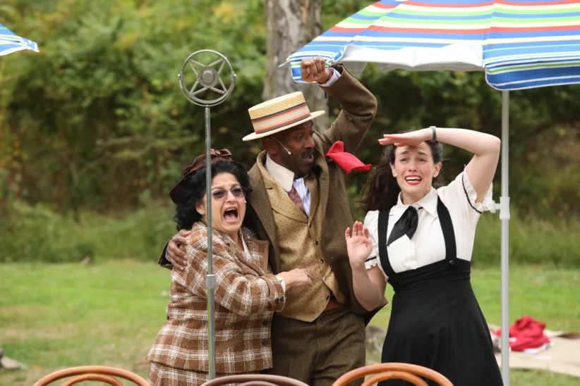 Three people dressed in vintage attire pose together outdoors by a retro microphone, under colorful umbrellas, during a theatrical performance.
