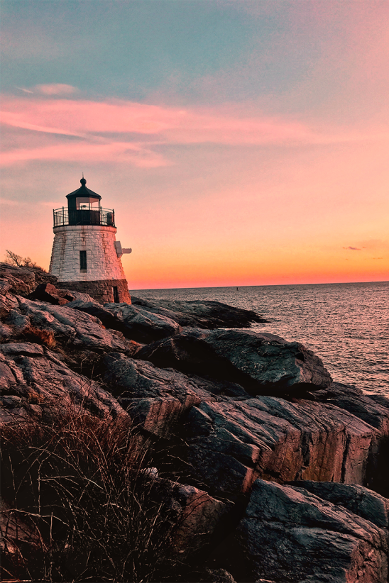 A lighthouse stands on rocky cliffs overlooking the ocean at sunset, with a pink-orange sky creating a tranquil coastal scene.