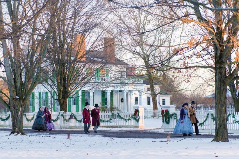 A snowy village scene featuring people in 19th-century attire walking near a white house with green shutters and festive garlands.