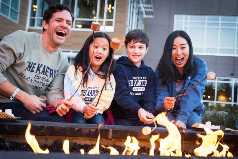 A family sits by a fire pit outside, roasting marshmallows on sticks while making s’mores, with a modern building in the background.