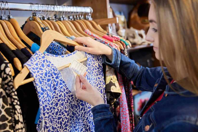 A woman in a denim jacket examines the tag of a blue patterned dress on a rack of clothing inside a brightly lit thrift store.