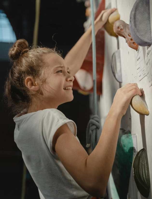 Young girl in a white shirt smiling while climbing an indoor rock wall, holding onto colorful handholds for support.
