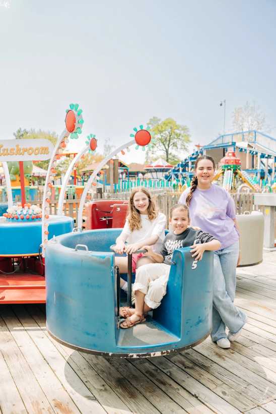 Three children enjoy a sunny day at an amusement park, sitting in a blue spinning ride, with colorful attractions in the background.