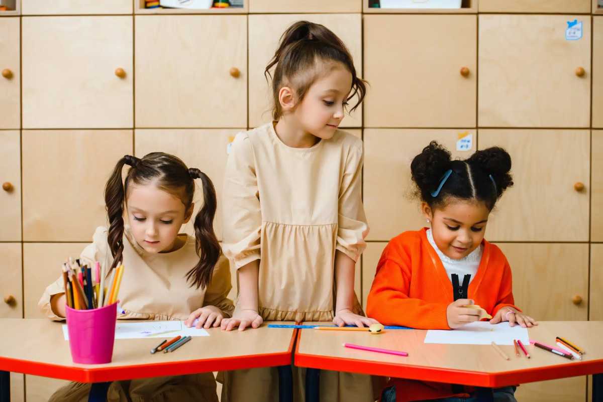 Three young girls draw at small tables in a classroom, using colored pencils, with storage cabinets neatly arranged behind them.