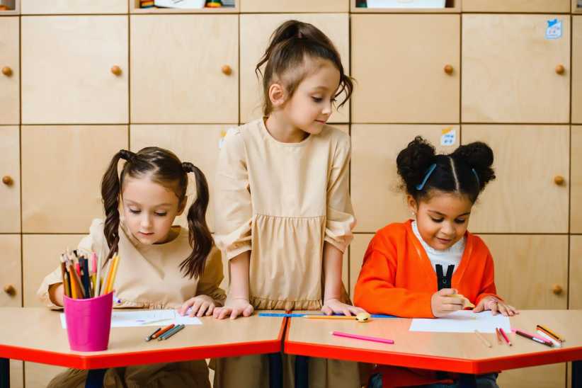 Three young girls draw at small tables in a classroom, using colored pencils, with storage cabinets neatly arranged behind them.