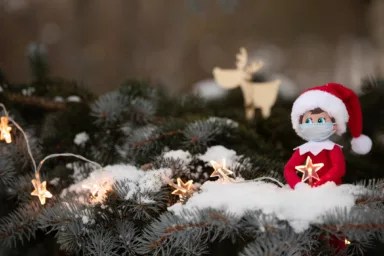A Christmas toy sits on the branches of a snow-covered Christmas tree in an embrace with a sparkling star during a pandemic. Coronavirus Covid Christmas: A festive toy doll wearing a real medical mask
