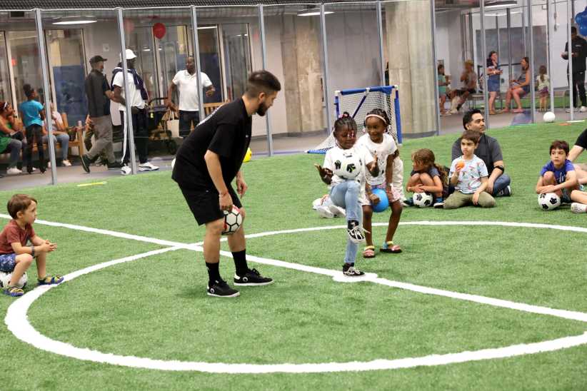 Children sit in a circle on indoor turf while a coach teaches soccer; two kids stand in the center, holding small soccer balls.