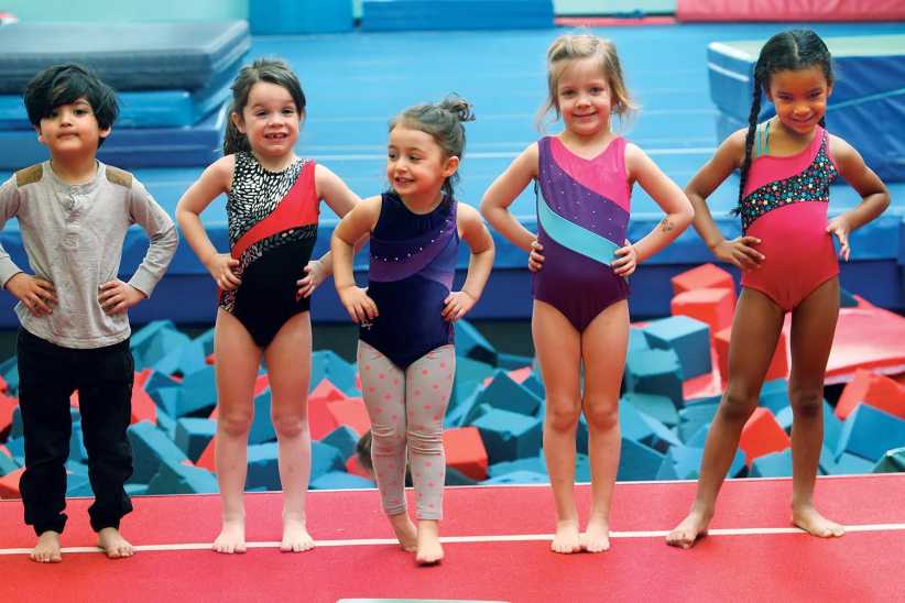 Five young children in gymnastics attire stand in a row with hands on hips, posing on a red mat with foam blocks behind them.