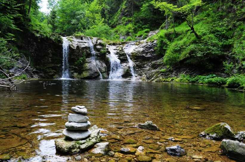 A tranquil forest scene features a small waterfall cascading into a clear pool, with a cairn of stacked stones on the rocky shore.