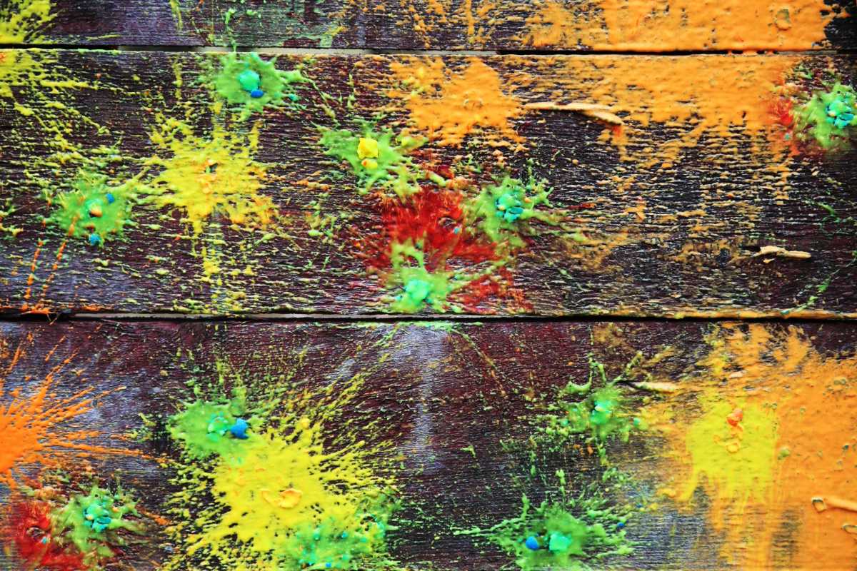 Colorful paintball splatters in yellow, orange, and green cover wooden boards, creating a vibrant, abstract burst effect on the surface.