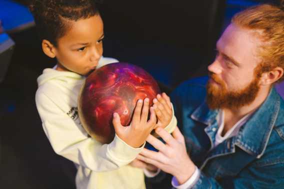 A young boy in a light-colored sweatshirt is being assisted by a bearded man in a denim jacket as he holds and prepares to bowl with a red and black bowling ball.