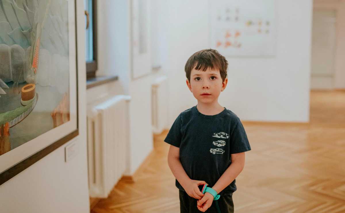Young boy stands in an art gallery wearing a navy blue t-shirt and turquoise wristwatch, gazing quietly at unseen artwork.