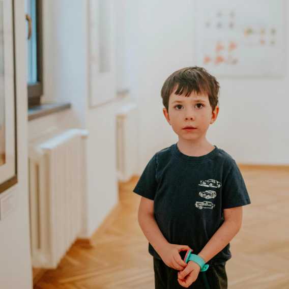Young boy stands in an art gallery wearing a navy blue t-shirt and turquoise wristwatch, gazing quietly at unseen artwork.