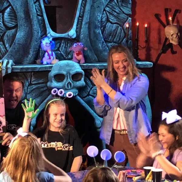 Children and an adult gather around a decorated table at a spooky-themed party, with monster props and skulls in a candle-lit setting.