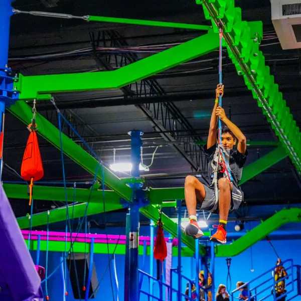 A child wearing a harness and helmet navigates an indoor ropes course, gripping a blue safety line among green and purple structures.