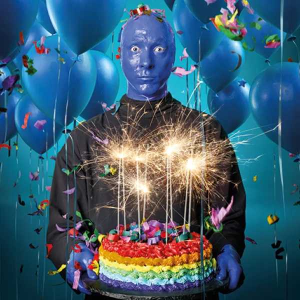 A person in blue gloves holds a vibrant rainbow cake with sparklers, surrounded by blue balloons and colorful confetti at a celebration.