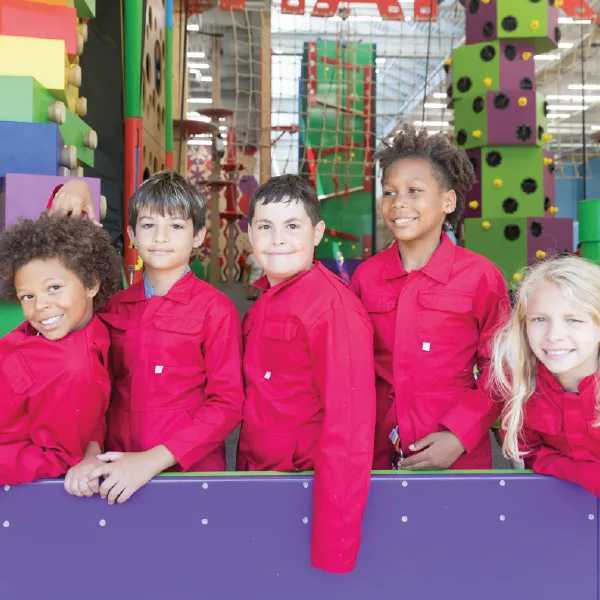 A group of children in matching red jumpsuits stands in front of a colorful indoor climbing wall, ready for a fun activity together.