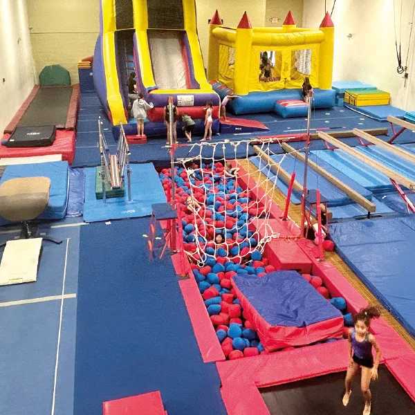 Children play in an indoor gym filled with bounce houses, a large inflatable slide, trampolines, and a foam pit with ropes and mats.