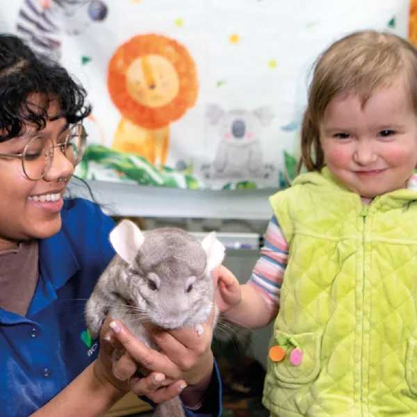 An adult and a child gently hold a chinchilla in front of a colorful animal-themed backdrop, engaging in an educational animal encounter.