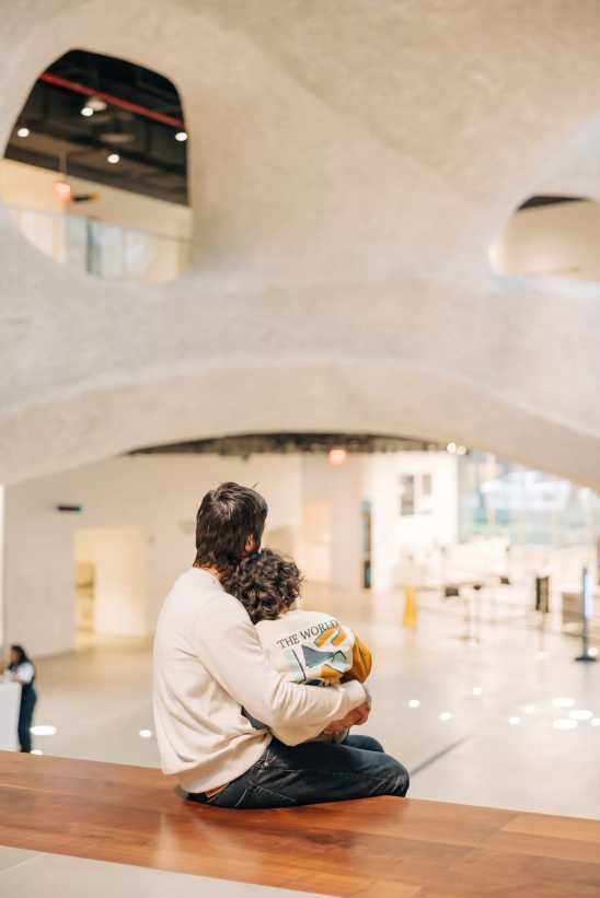 An adult and child holding hands walk through a modern museum with wooden ceilings, plant displays, and interactive exhibits.