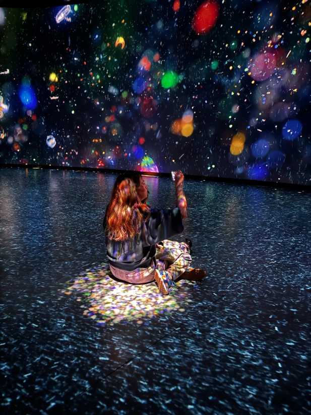 A person sits on a floor illuminated by projected colorful lights, surrounded by a vivid, immersive digital art display.