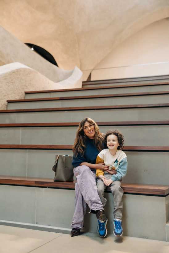 A woman and child sit smiling on wide wooden steps in a modern space with light-colored stone walls and an arched ceiling.