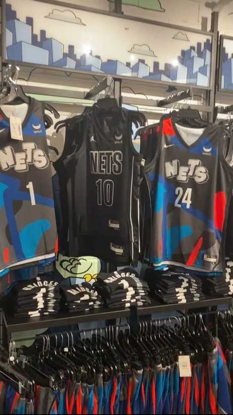 Brooklyn Nets basketball jerseys in various colors and numbers are displayed on hangers and folded on shelves in a merchandise store.