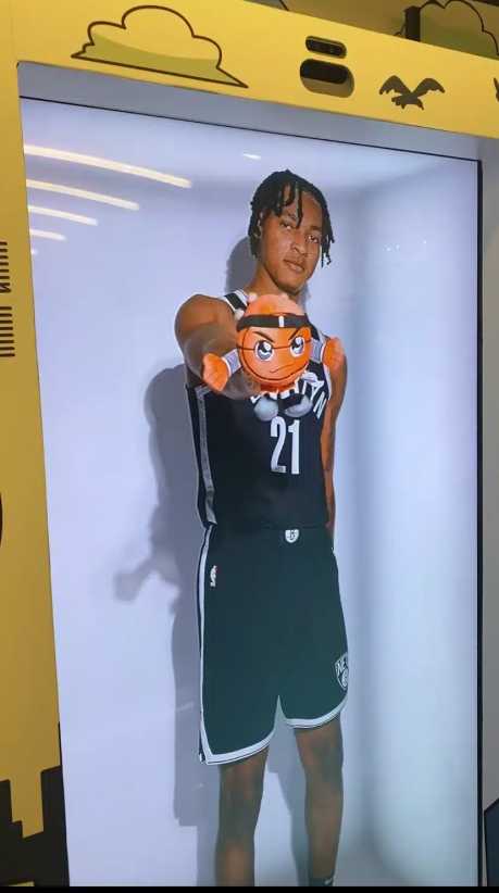 A person in a Brooklyn Nets basketball uniform stands inside a yellow display, holding a plush toy with an expressive face pointed forward.