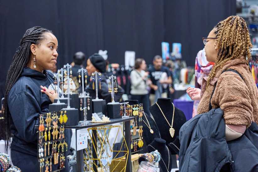 Two women converse at a jewelry booth during an indoor holiday market, surrounded by handmade earrings, necklaces, and shoppers.