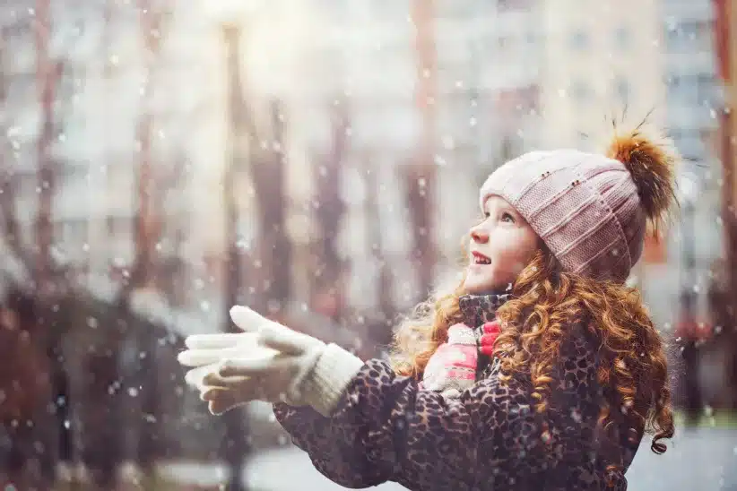 Little girl stretches her hand to catch falling snowflakes.