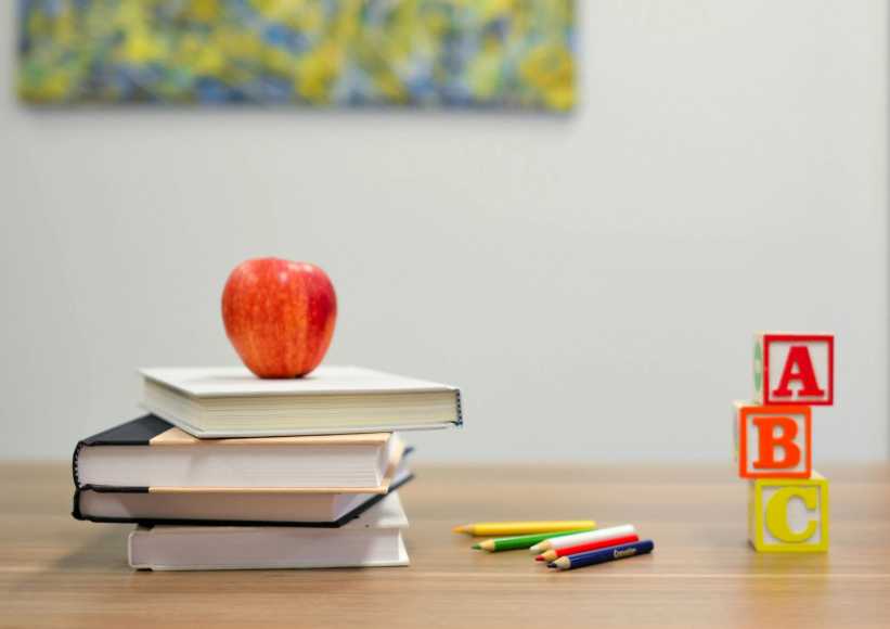 Red apple on stacked books with colored pencils and ABC letter blocks on a wooden desk in a classroom setting.