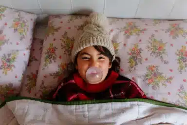 Girl wearing beige warm hat making bubble lying in the bed under blanket