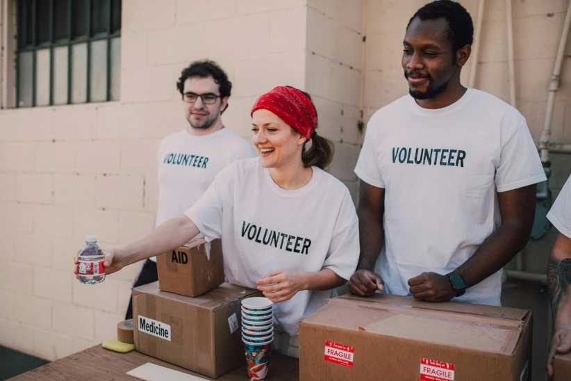 Three smiling volunteers in white "VOLUNTEER" t-shirts stand behind a table outdoors, handing out water bottles, medicine boxes, and cups during a community aid event.