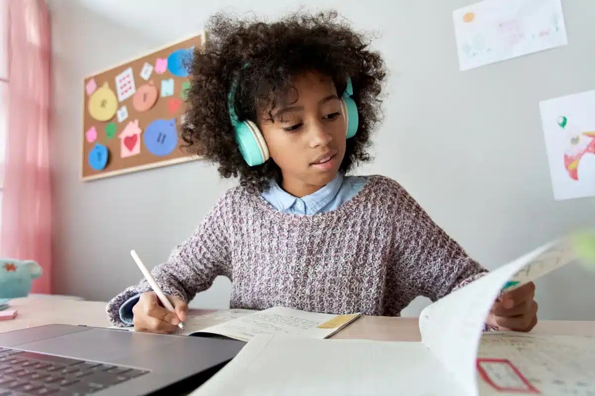 Smart african mixed race kid girl wearing headphones listening lesson learning online computer doing homework at home. Cute black child studying distance school education writing notes sitting at desk