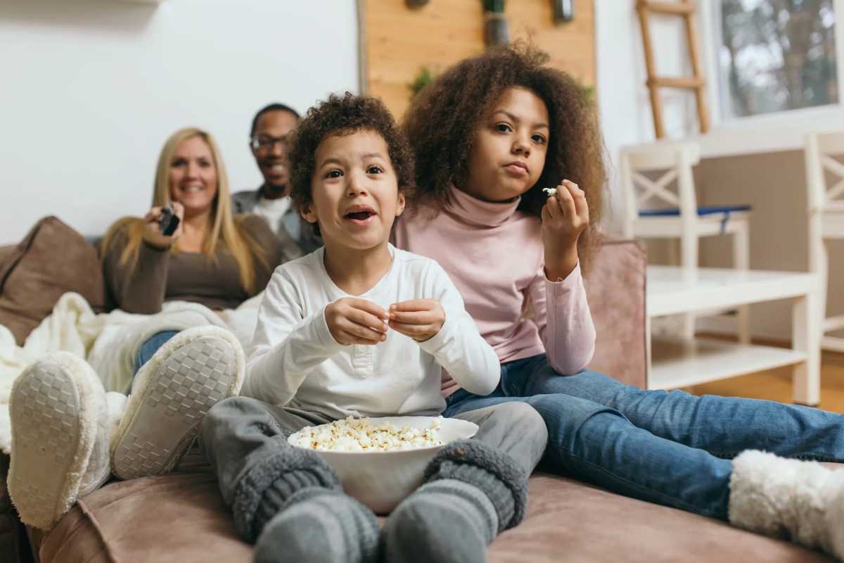 Family sitting on a couch watching TV together, two kids in front eating popcorn, parents smiling in the background.