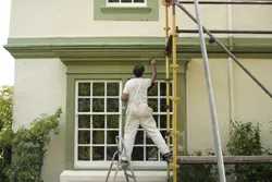 House painter standing on scaffolding and ladder while painting the exterior wall of a residential home.