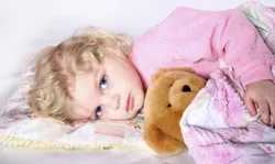 A young blonde girl with blue eyes sleeps peacefully in bed, hugging a teddy bear, wrapped in a pink blanket on white sheets.