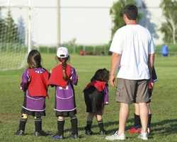 A man in a white t-shirt stands on a grassy field with young soccer players in purple and red jerseys.