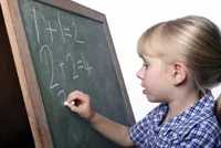 A young girl with blonde hair writing simple addition equations on a green chalkboard with white chalk.