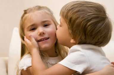 A young boy in a white t-shirt gently kissing a smiling young girl's cheek while hugging her.