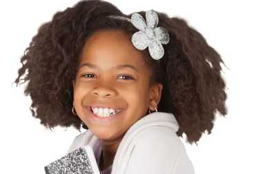 Smiling young Black girl with voluminous natural hair and a silver flower clip, holding a black and white composition notebook.