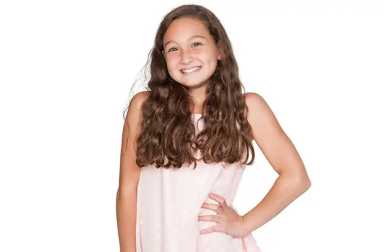 A young girl with long, wavy brown hair smiling while posing with one hand on her hip against a white background.