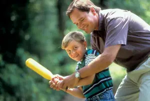 A man smiling as he helps a young boy in a striped shirt hold and swing a yellow plastic baseball bat.
