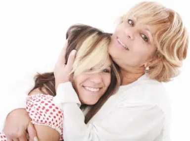 A blonde woman in a white shirt warmly embraces a younger girl with dark hair and blonde highlights against a white background.