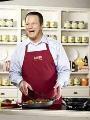 A man in a red apron labeled "David" smiles while cooking food in a skillet on a stovetop in a kitchen.