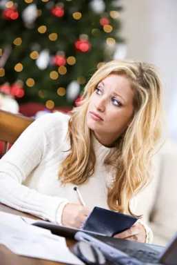 A blonde woman looks pensively away while writing in a notebook, with a blurred Christmas tree in the background.