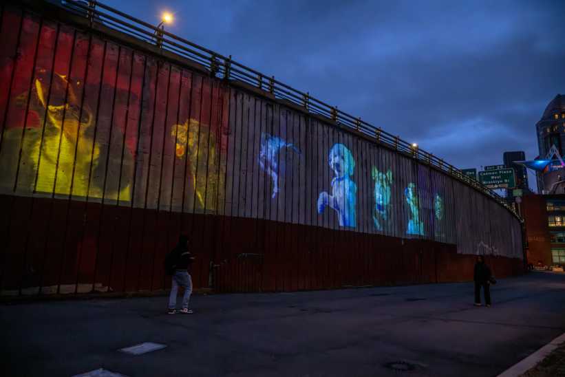 Illuminated video art projections of animals and people light up a curved Dumbo wall at night, as viewers watch the display.