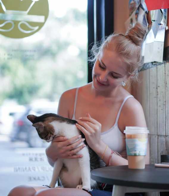 A woman with blonde hair in a bun wearing a white tank top sits by a window holding a tabby cat. A coffee cup is on the table nearby.