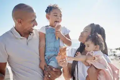 Black family, beach and ice cream with children and parents on sand by the sea or ocean during summer together. Kids, travel and relax with a mother, father and daughter siblings bonding in nature