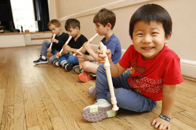 Smiling child in red shirt holds a recorder while sitting on the floor with other kids playing recorders in a music class.
